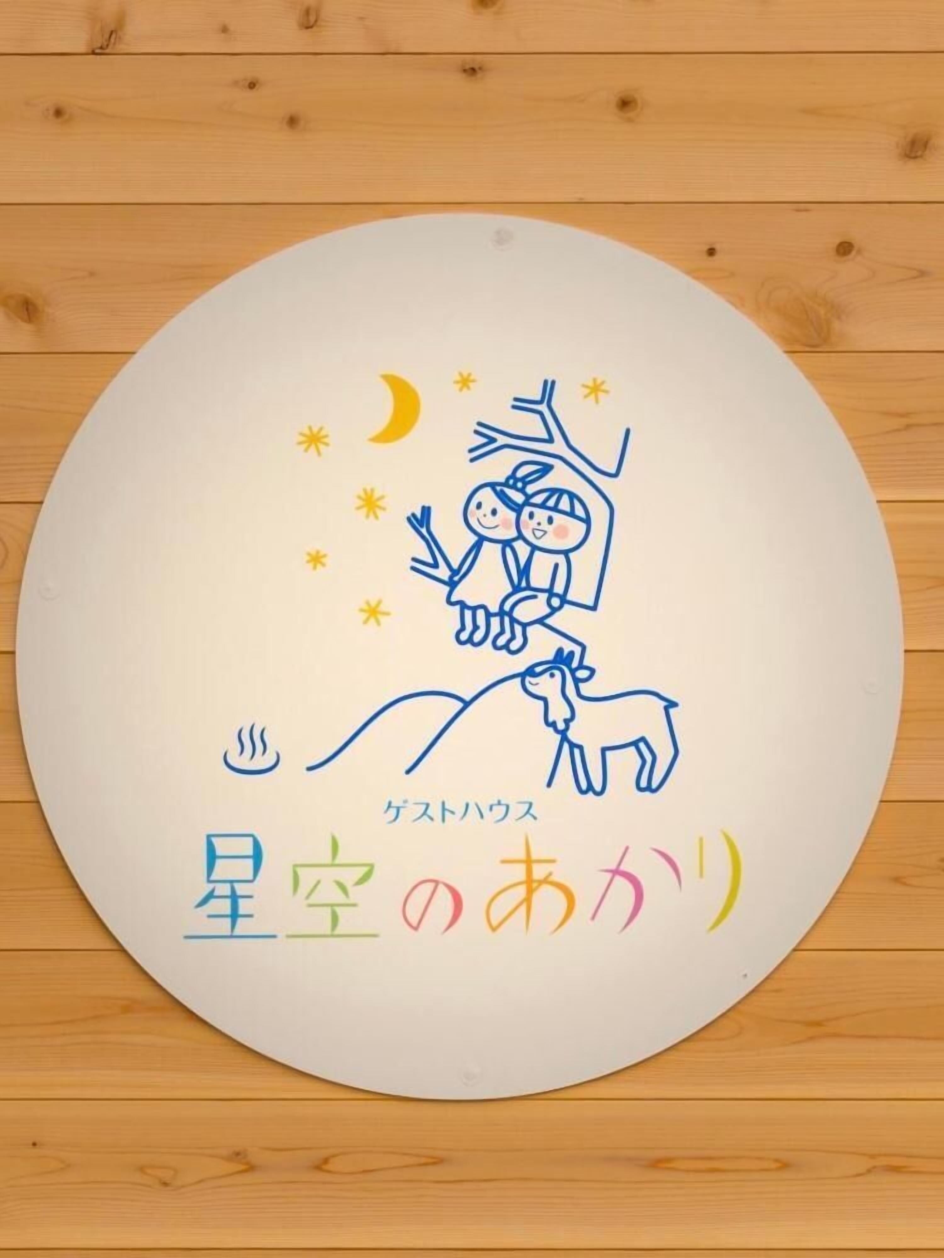 Un letrero redondo y blanco con un dibujo sencillo de dos niños en una rama de árbol, una luna creciente, estrellas y texto en japonés.