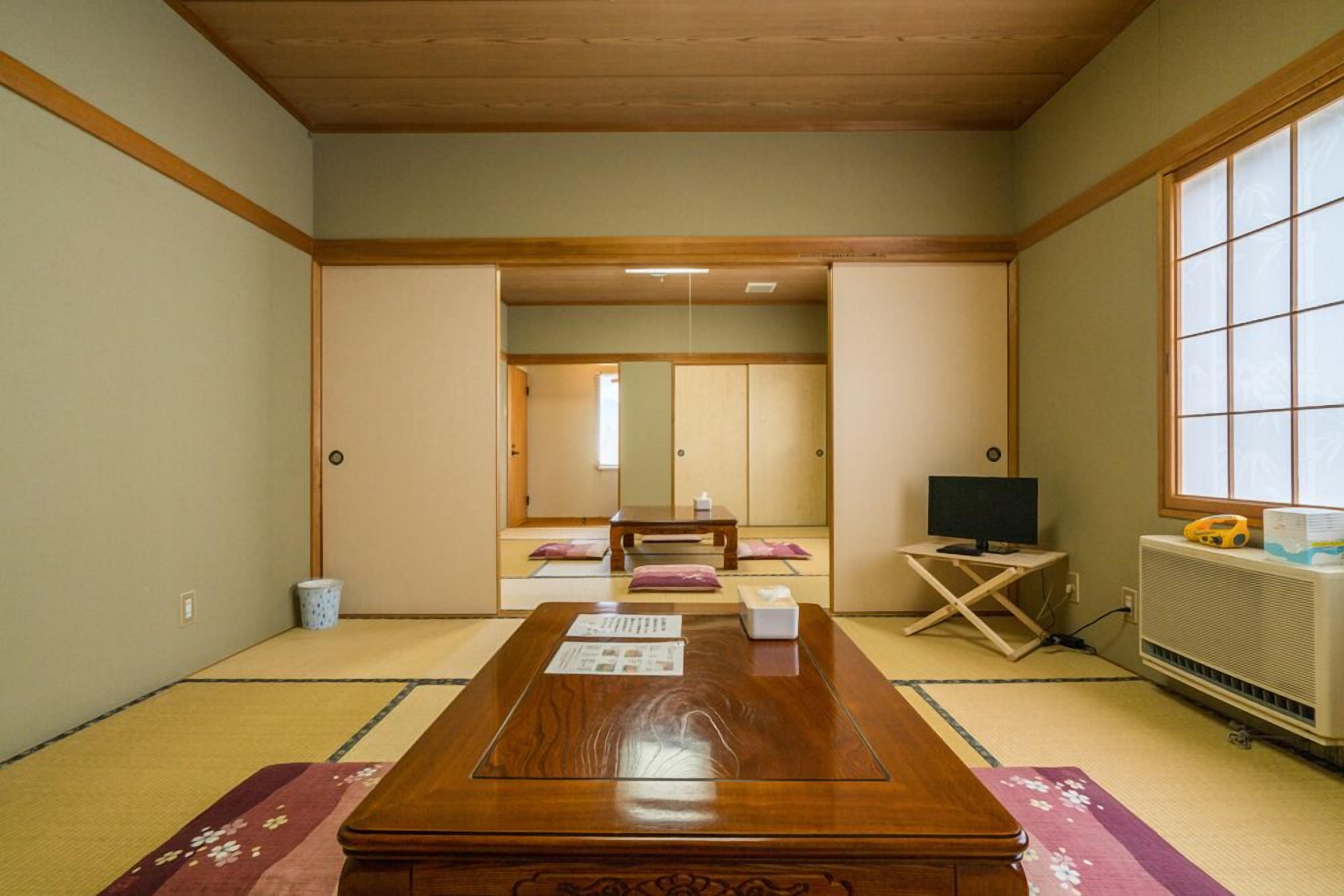 A traditional Japanese room with tatami flooring, a low wooden table, a television, and a futon bed in the background.