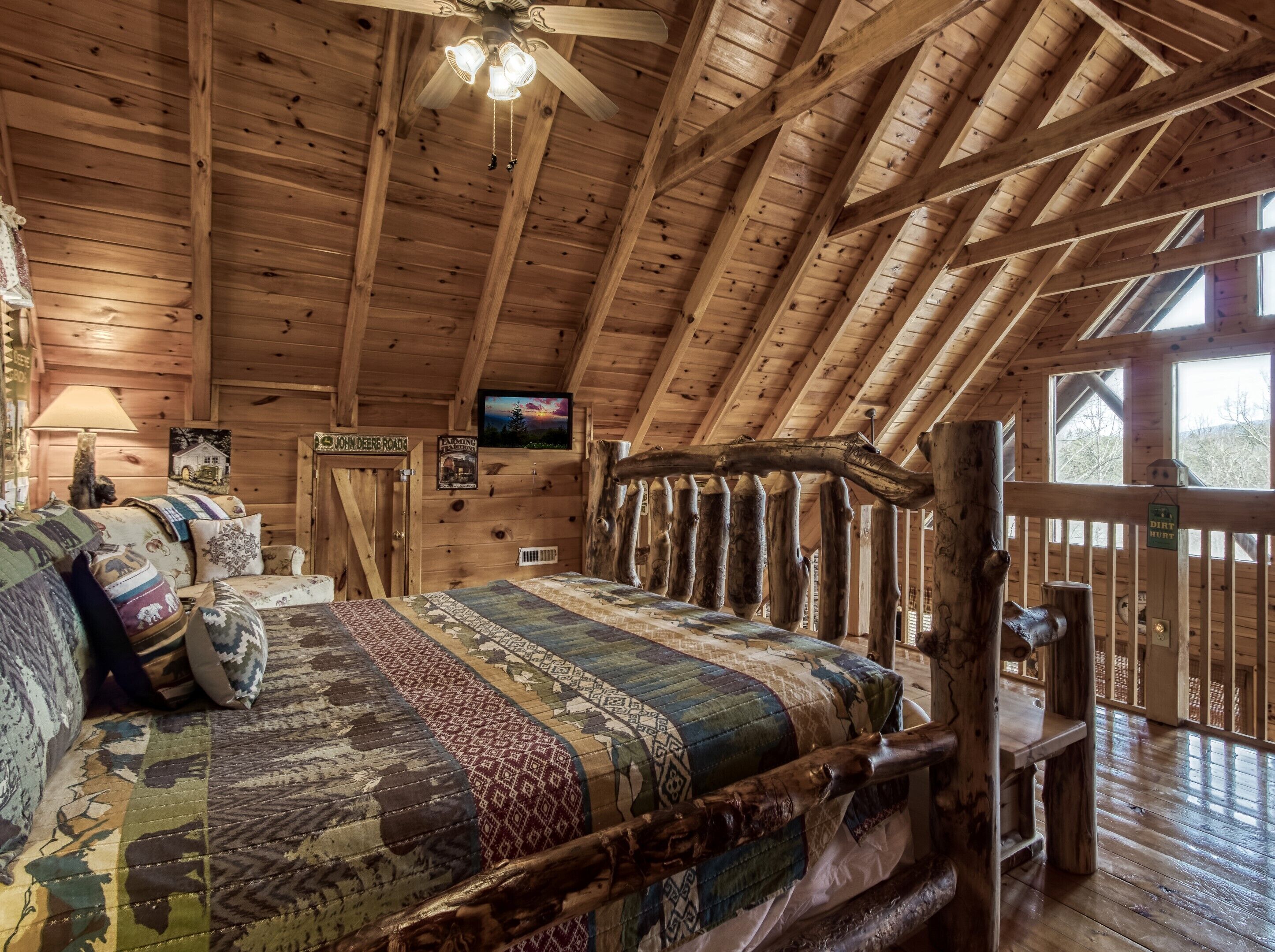 A cozy log cabin interior with a wooden ceiling, a bed with patterned bedding, a television on the wall, and a sign indicating a dirt hut.