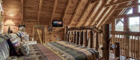 A cozy log cabin interior with a wooden ceiling, a bed with patterned bedding, a television on the wall, and a sign indicating a dirt hut.