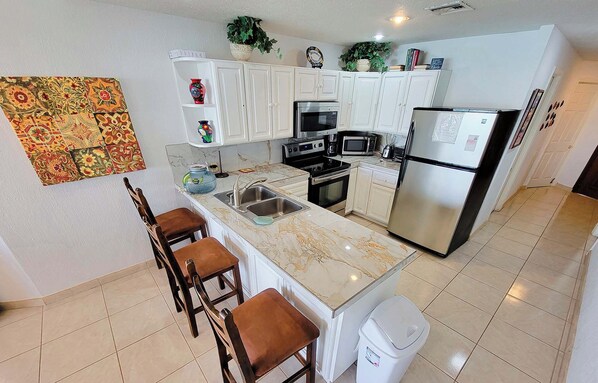 A kitchen with white cabinets, a marble countertop, a sink, a stove, a microwave, a refrigerator, and a trash can.