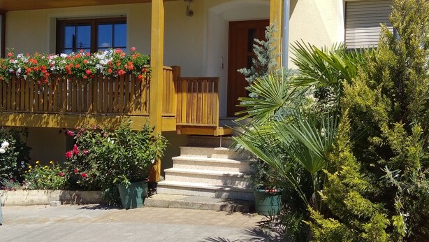 Una casa con balcón de madera, una escalera que lleva a la entrada y varias macetas con plantas alrededor de la propiedad.