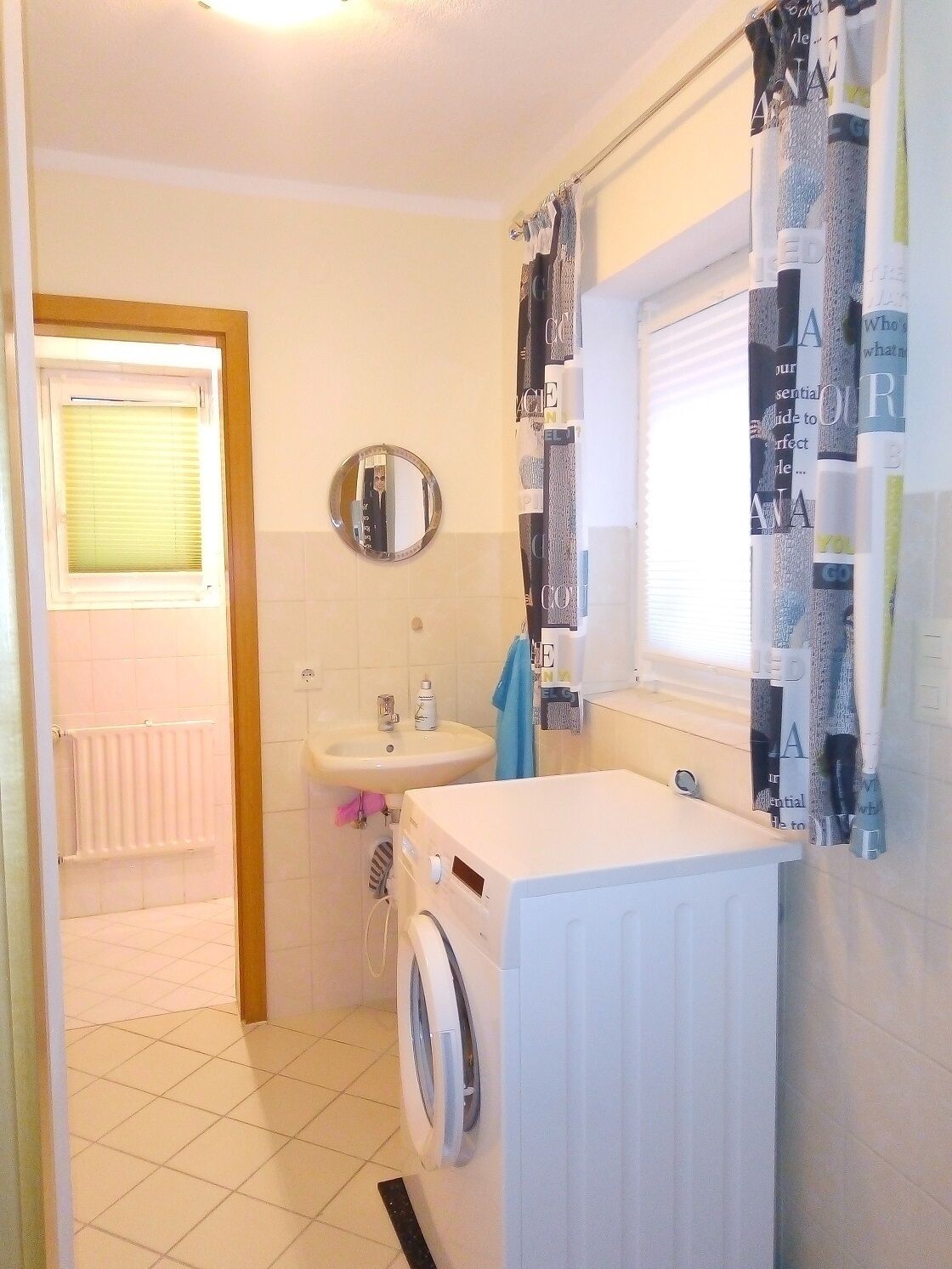 A compact bathroom with a washing machine, a sink, a mirror, and a window with a curtain.