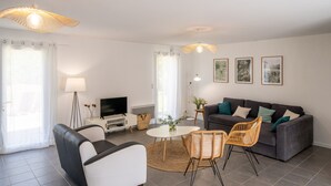 A modern living room with a grey sofa, white armchair, wicker chairs, a small round table, and a TV on a white stand. - Comfortable cottage in peace between sea and marsh! (Sallertaine)
