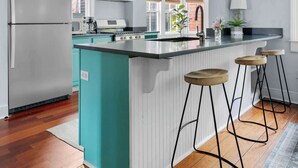 A modern kitchen with teal cabinets, a stainless steel refrigerator, a central island with stools, and a wooden floor. - Charleston Riviera | King Street Home - Walkable! (Charleston)