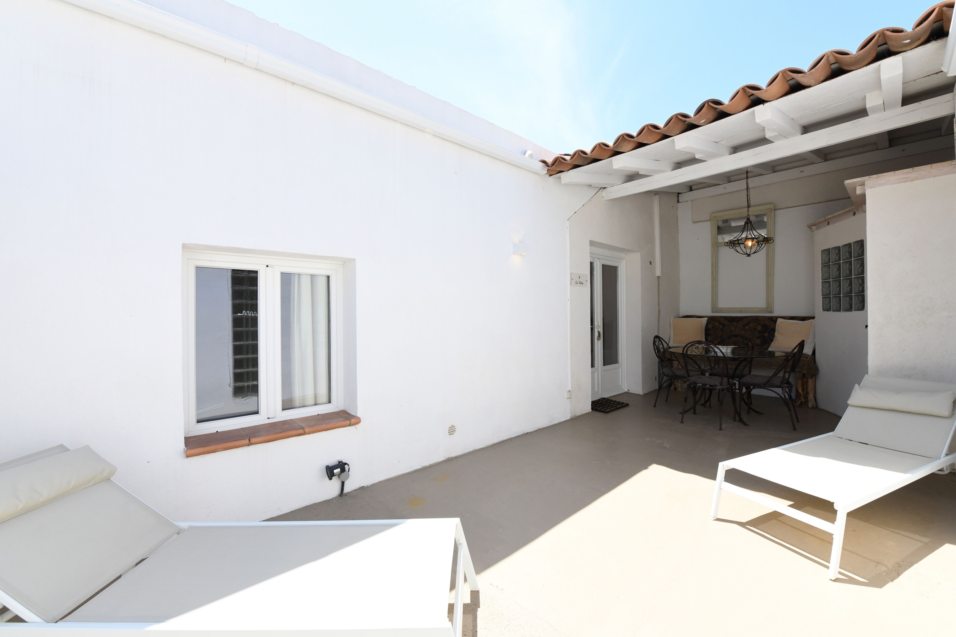 Un bâtiment blanc avec un toit recouvert de tuiles, une fenêtre, une porte, un coin repas avec une table et des chaises, et deux fauteuils de salon blancs.