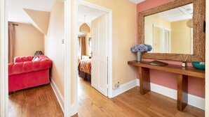 A hallway with a red upholstered bench, a wooden console table with a mirror, and a door leading to a bedroom. - Lovely 2BR, Richmond Hill & Parking (Richmond upon Thames)