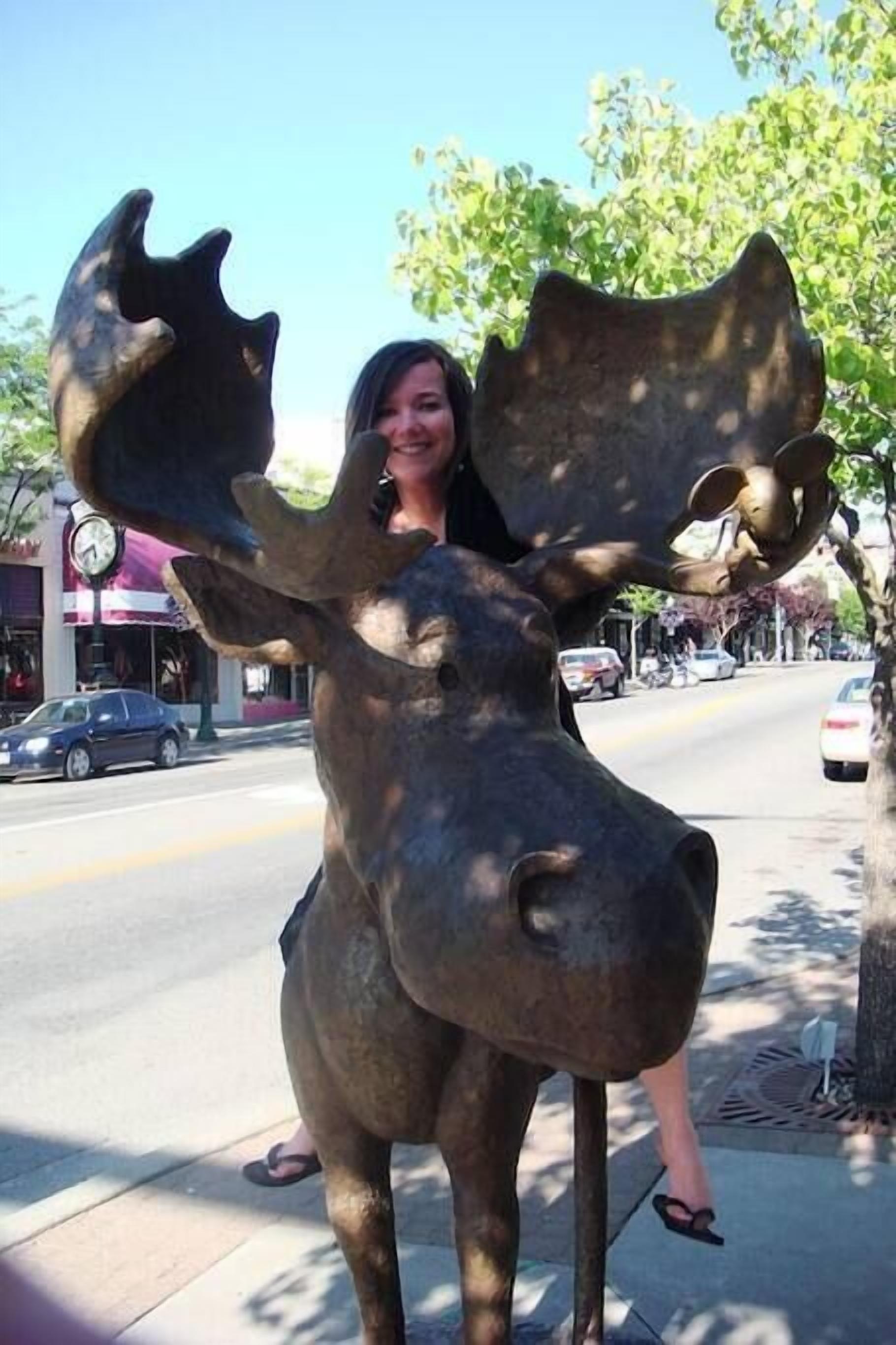 A person posing with a moose statue on a sidewalk.