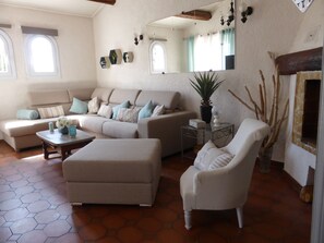 Een woonkamer met een beige bank, een witte fauteuil, een salontafel en een bijzettafeltje met een plant. Aan de muur hangt een grote spiegel en er is een raam met gordijnen.