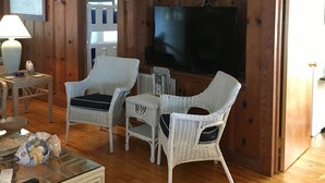 Ein Zimmer mit Holzwänden, einem Fernseher, Korbmöbeln und einem Glastisch.