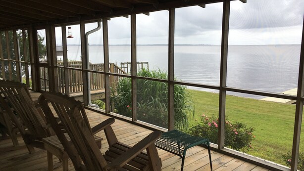 Eine überdachte Terrasse mit Holzstühlen und einer Bank, mit Blick auf eine Rasenfläche und das Meer.