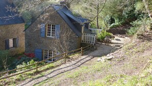 A stone house with a sloped roof, a wooden deck, and a gravel path leading to a stone wall. - The home of the Moulin de Kérangoc (Melgven)