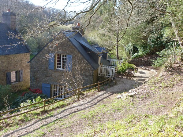 A stone house with a sloped roof, a wooden deck, and a gravel pathway leading to a stone wall.