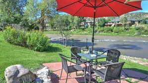 Een terras buiten met een tafel en stoelen, een rode parasol, en een uitzicht op een rivier en huizen op de achtergrond.