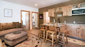 Een gezellige woonkamer met een bank, voetenbankje en een eethoek met houten stoelen en een tafel. Er is een keuken met houten kastjes en een magnetron.
