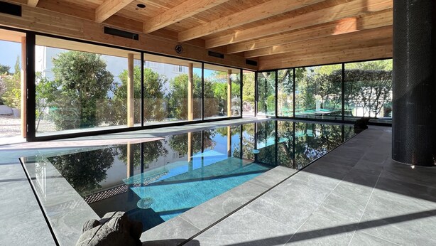 Ein moderner, überdachter Pool mit Glaswänden und Holzbalken, umgeben von einem Garten.