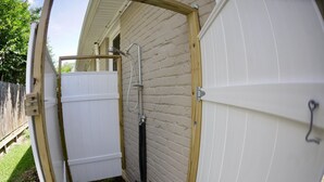 A compact outdoor shower area with a wooden frame, a metal showerhead, and a white tiled wall.