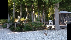 Un espace recouvert de gravier avec une terrasse en bois, entouré d’arbres et comprenant un coin salon abrité avec une chaise.