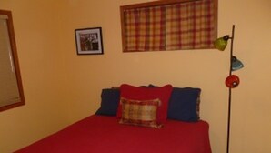 Een slaapkamer met een rode plaid, twee kussens, een vloerlamp, een geruit gordijn en een ingelijste foto aan de muur.