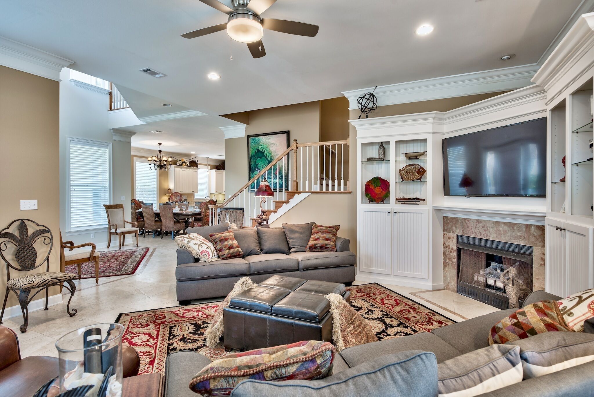 A spacious living room with a grey sofa, ottoman, and a fireplace. There is a dining area in the background with a staircase leading to an upper level.