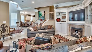 A spacious living room with a grey sofa, ottoman, and a fireplace. There is a dining area in the background with a staircase leading to an upper level. - Bird of Paradise, see the # of 5-stars-Just steps from Beach (Miramar Beach)