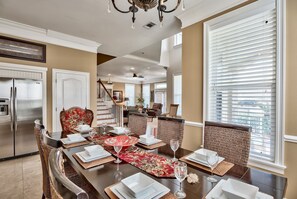A dining area with a wooden table set for six, surrounded by wicker chairs, a red floral runner, and a view of a living room in the background.