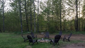 A campsite with four Adirondack chairs arranged around a central fire pit, surrounded by a grassy area and a dirt path, with tall trees in the background. - In The Heart Of The Hiawatha National Forest ~ Easy Access To The Trail System (Shingleton)