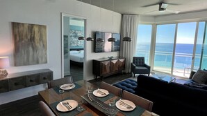 A modern hotel room with a dining area, a living room with a view of the ocean, and a large mirror reflecting the room.