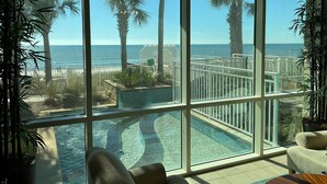 A view of a swimming pool with a curved design, surrounded by a white fence and palm trees, with a clear view of the ocean beyond.