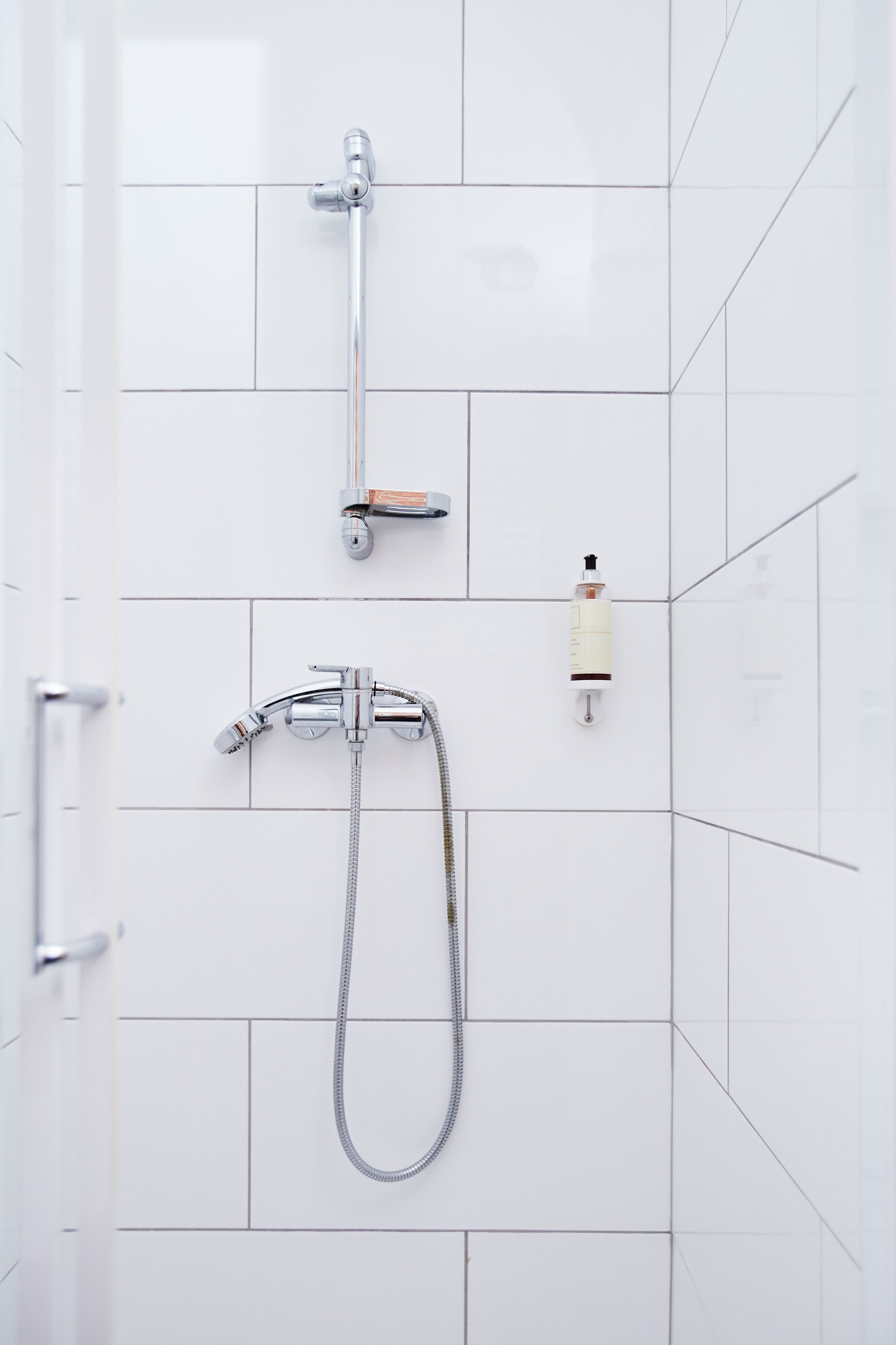 A bathroom with white tiled walls, a shower with a handheld showerhead, and a bottle of shampoo on the wall.