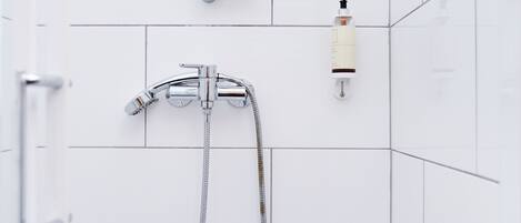 A bathroom with white tiled walls, a shower with a handheld showerhead, and a bottle of shampoo on the wall.