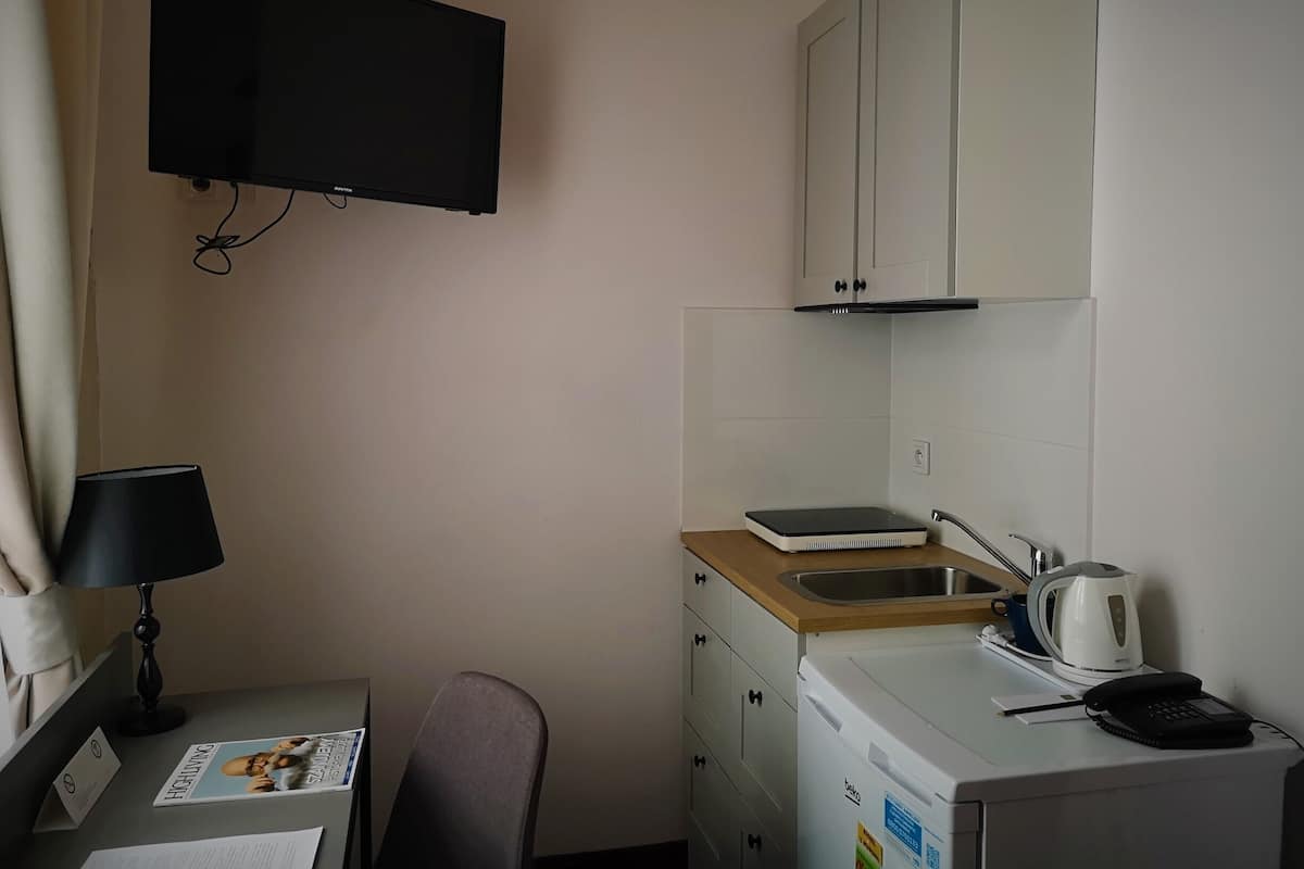 A compact hotel room with a flat-screen TV mounted on the wall, a desk with a lamp, a chair, a small kitchenette with a sink and a kettle, and a minibar.