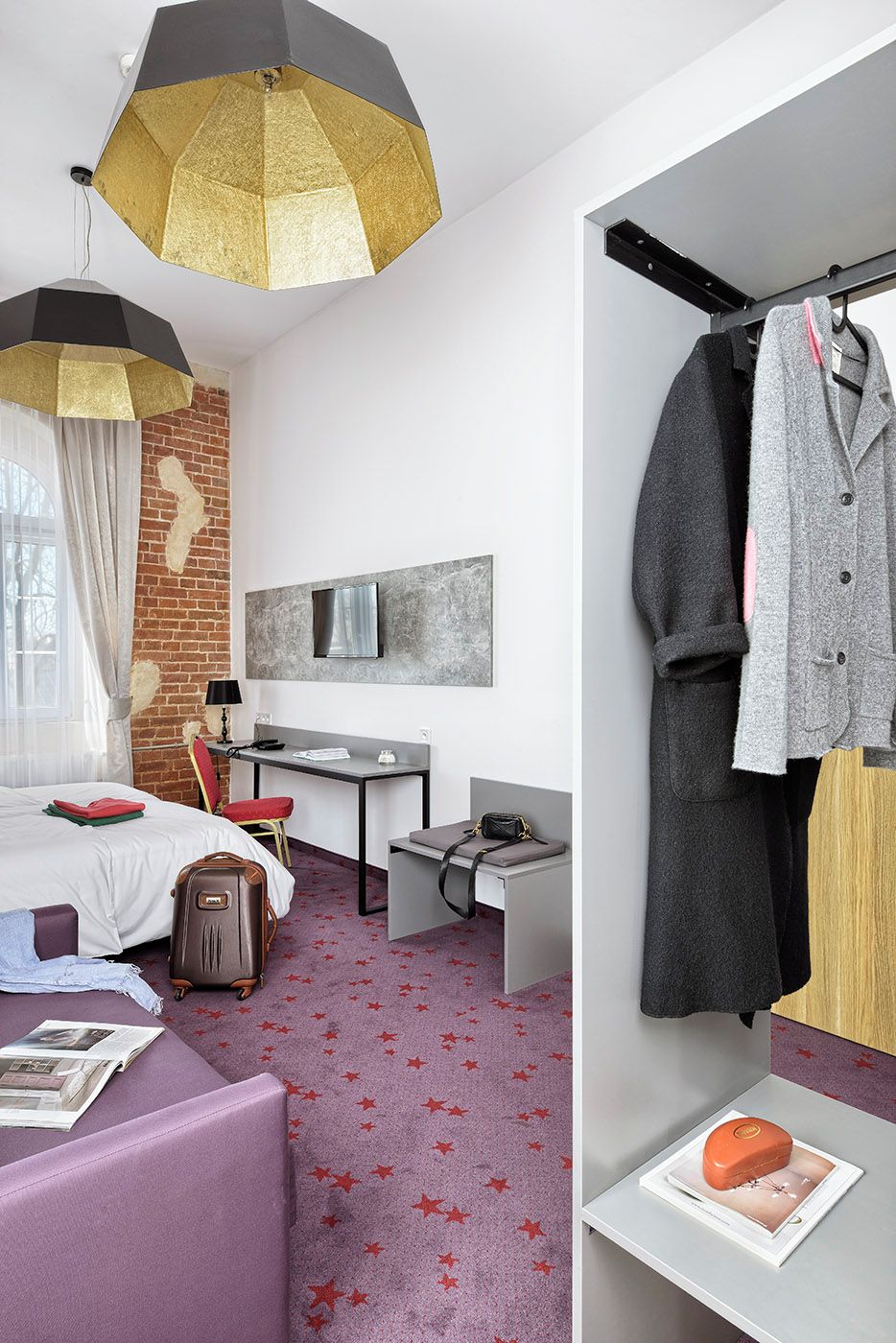 A hotel room with a bed, desk, chair, and a coat rack with clothes.