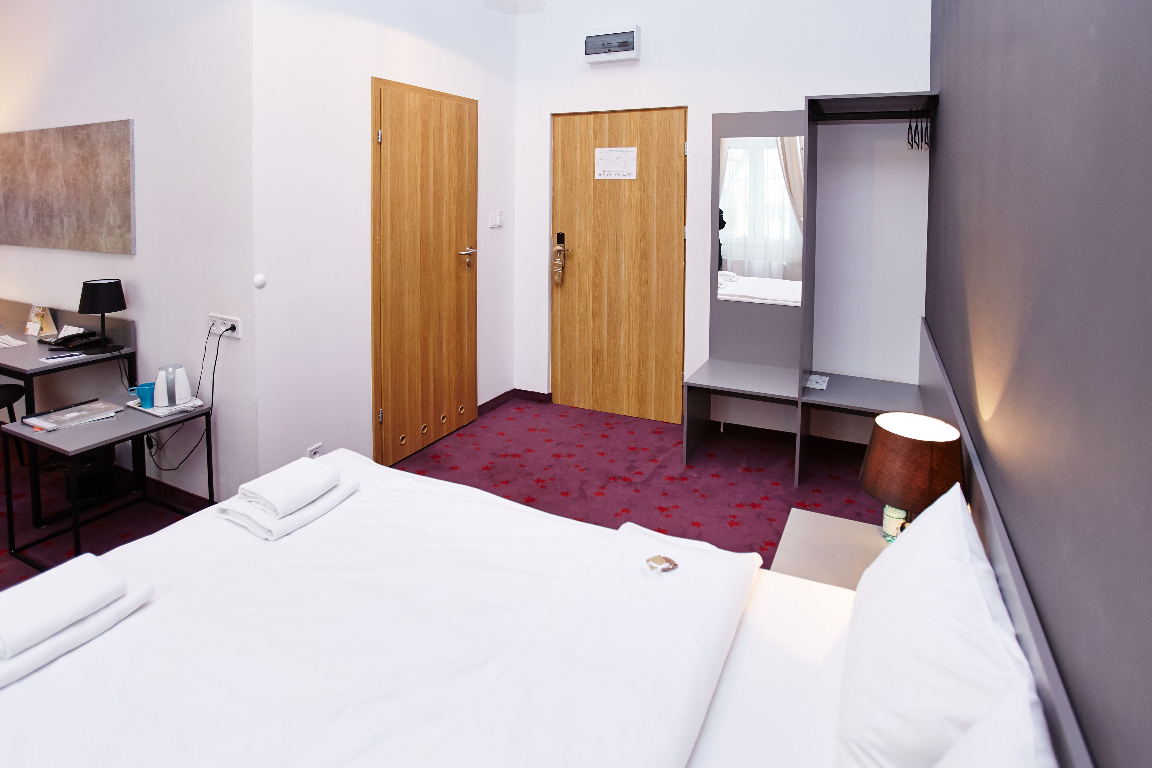 Habitación de hotel con cama, escritorio con lámpara, silla, armario y espejo.