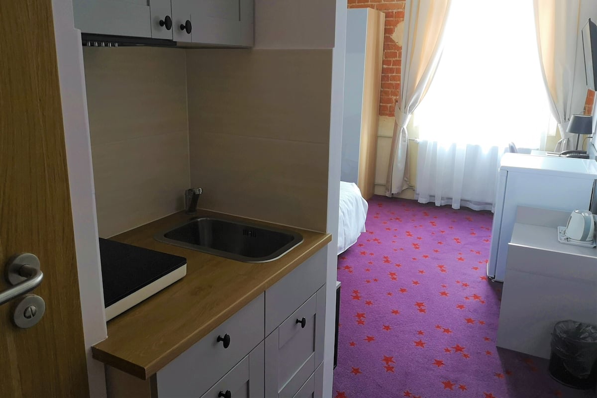 A compact hotel room with a kitchenette, a bed, a desk, and a window with curtains.