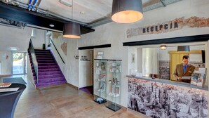 A reception area with a reception desk, a man standing behind it, a staircase, and a wall with a historical mural. - Koszary Arche Hotel (Gora Kalwaria)