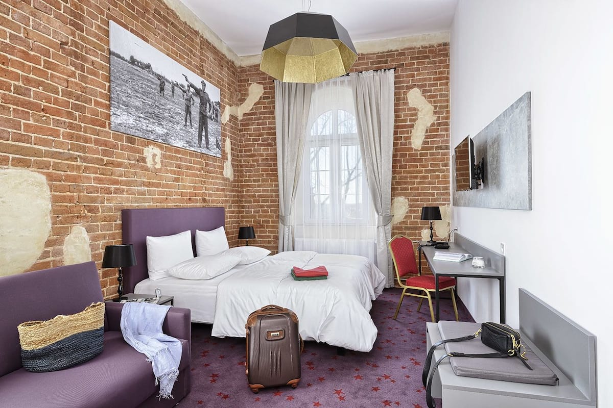 A hotel room with a brick wall, a bed with white bedding, a purple headboard, a desk with a chair, a suitcase, and a painting on the wall.