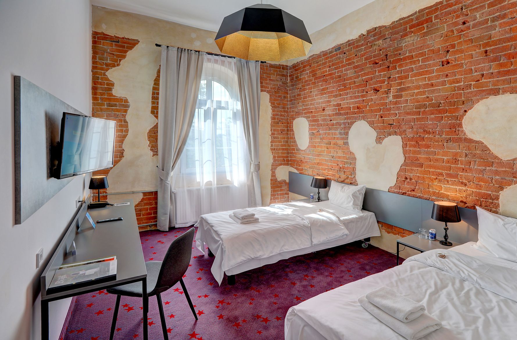 Habitación de hotel con pared de ladrillo, cama, escritorio con silla, televisor y ventana con cortinas.