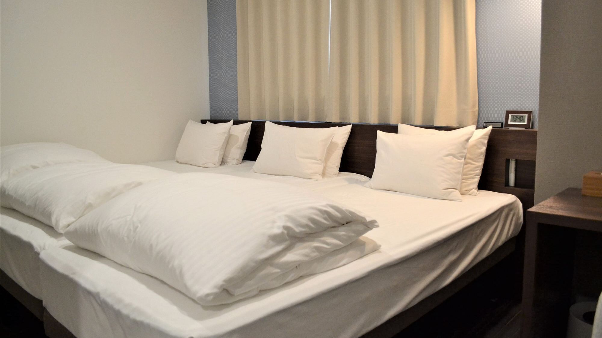 A neatly made bed with white linens and pillows, a wooden headboard, a nightstand with a clock, and a window with curtains.