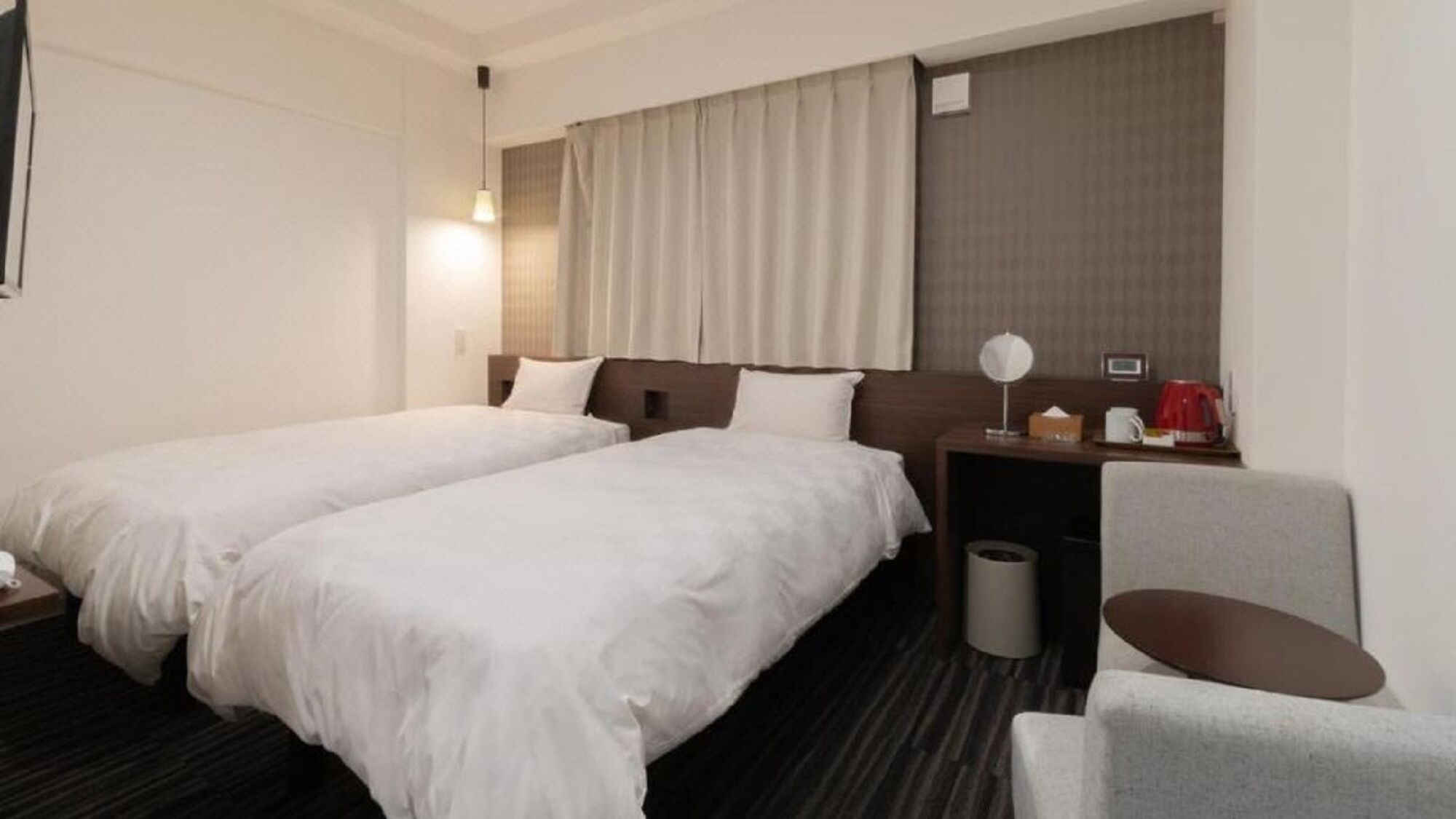 A hotel room with two beds, a desk, a chair, a lamp, a trash bin, and a window with curtains.