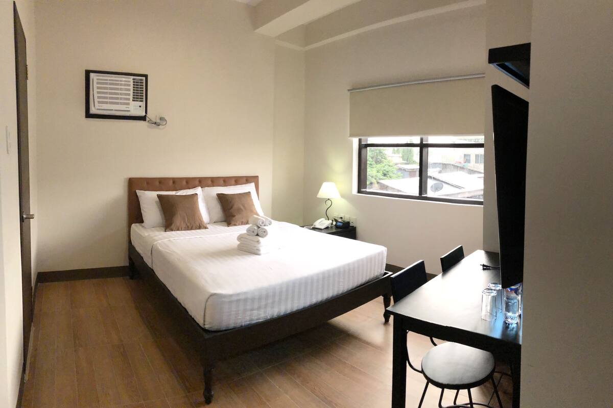 A hotel room with a bed, a desk, a chair, a lamp, and a window.