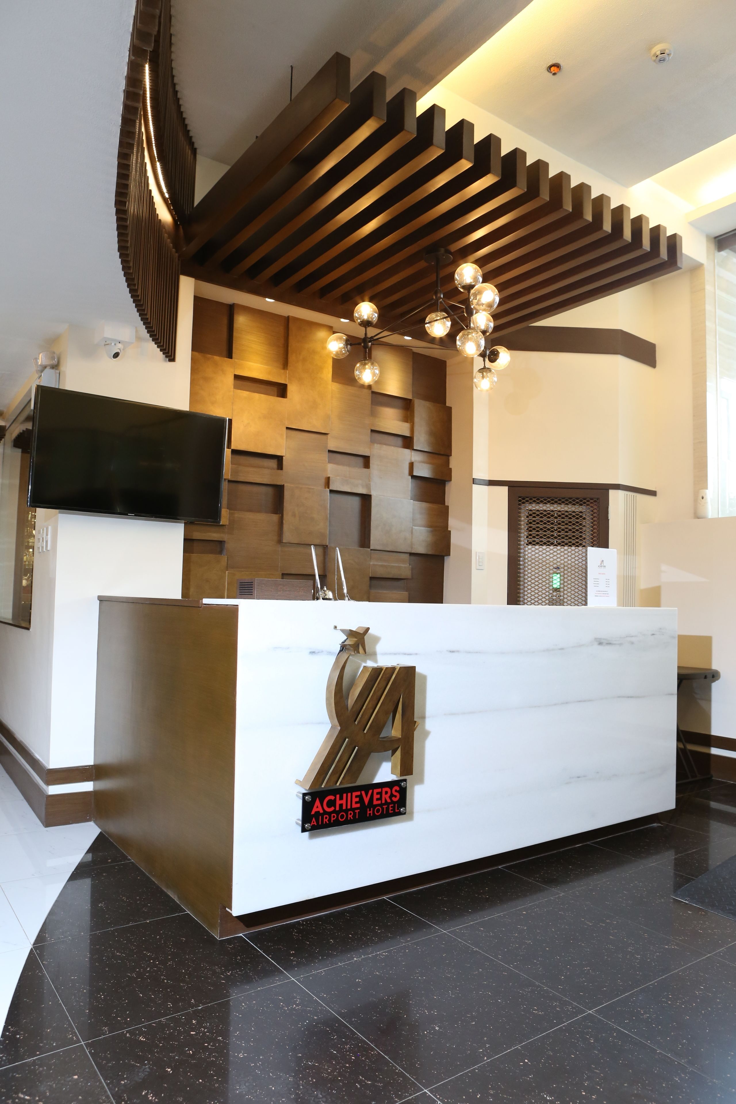 A reception desk with a modern design, a flat-screen TV, and a sign reading "Achievers Airport Hotel."