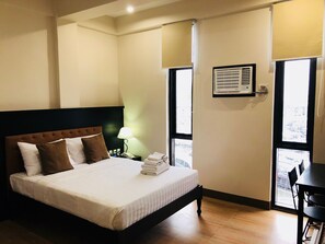 A hotel room with a bed, bedside table, lamp, air conditioning unit, and a view through the window. - Achievers Airport Hotel (Pasay)