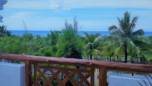 Une balustrade en bois surplombe un paysage tropical, avec des palmiers et une vue sur l’océan.