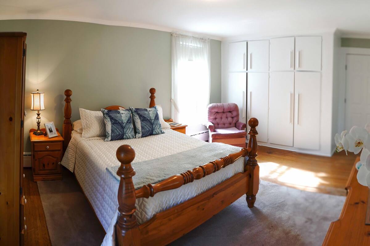 A bedroom with a wooden four-poster bed, a nightstand with a lamp, a chair, a wardrobe, and a mirror.