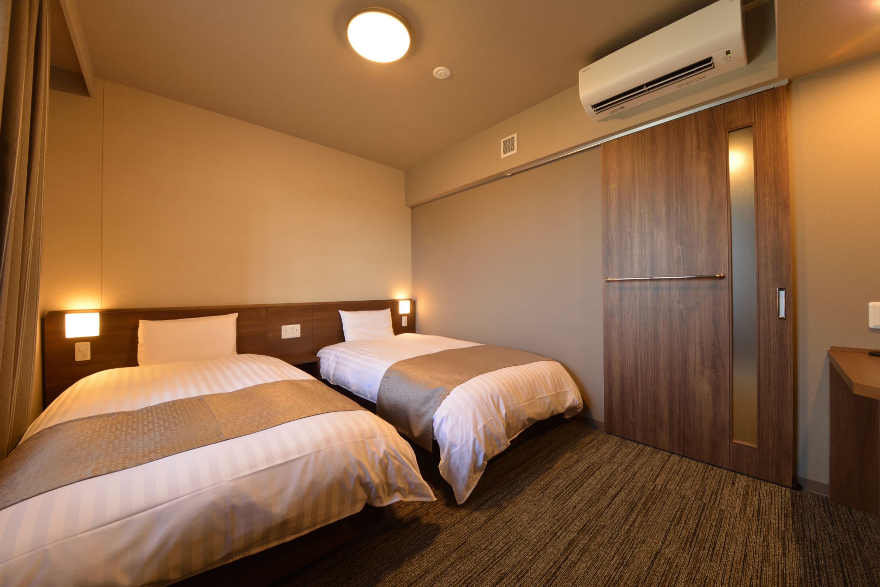 A hotel room with two beds, a wooden wardrobe, and an air conditioning unit.