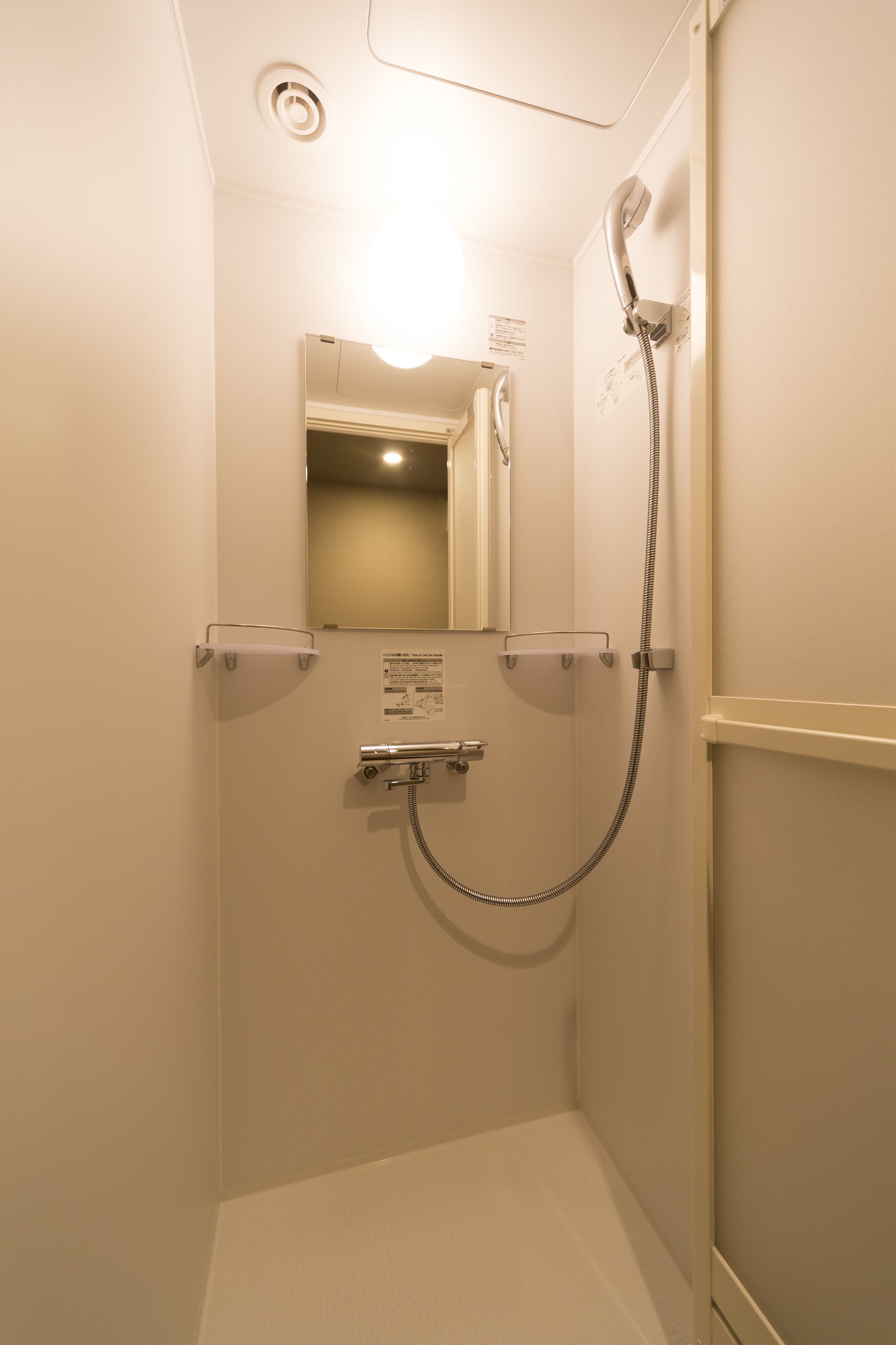 A small, enclosed shower stall with a handheld showerhead, a wall-mounted mirror, and a ceiling light.