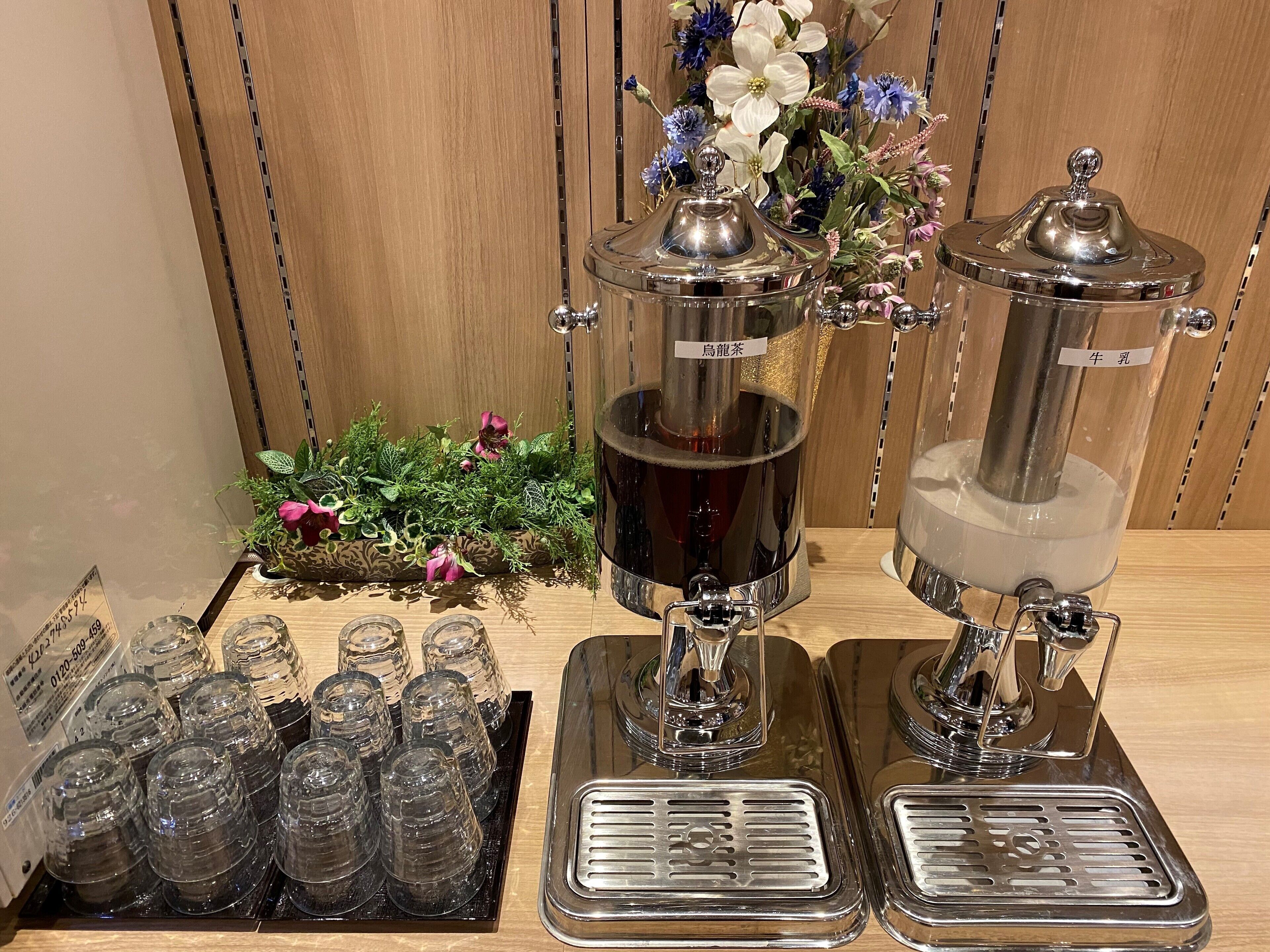 A coffee station with two coffee dispensers, a tray of clear glass cups, and a decorative plant.
