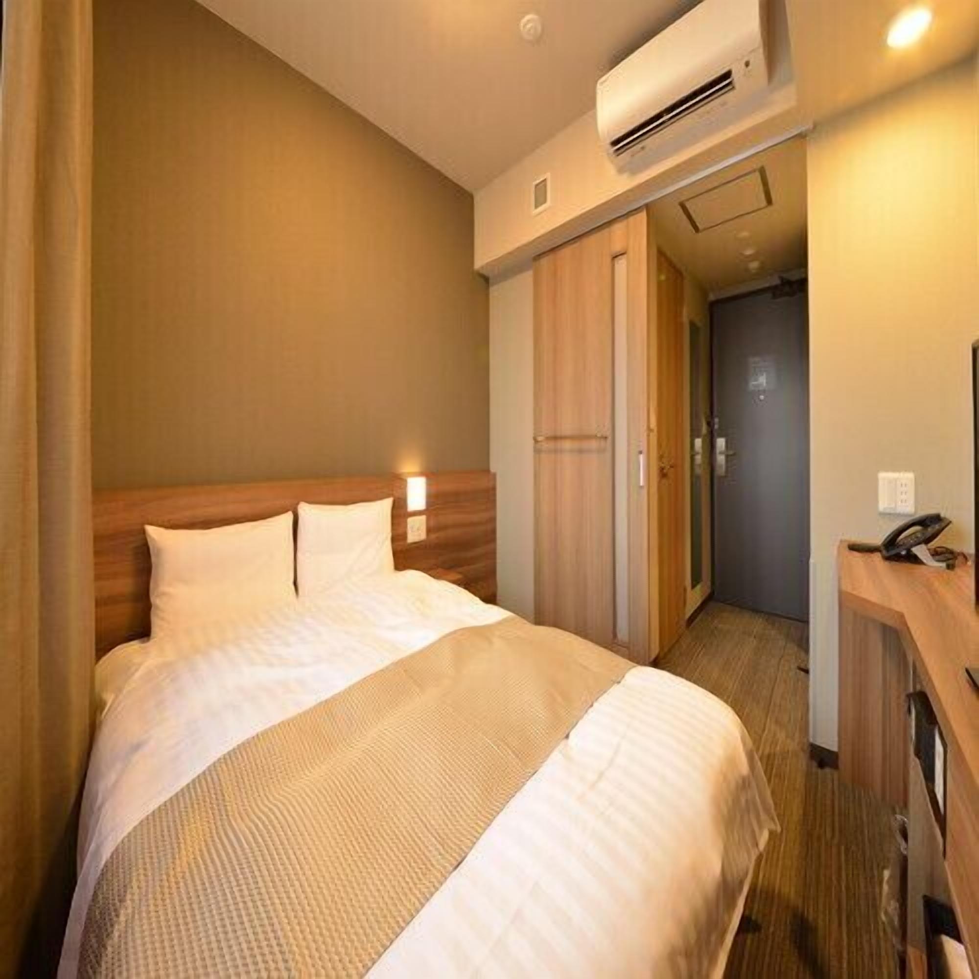 A hotel room with a bed, an air conditioning unit, a desk with a phone, and a door leading to another room.