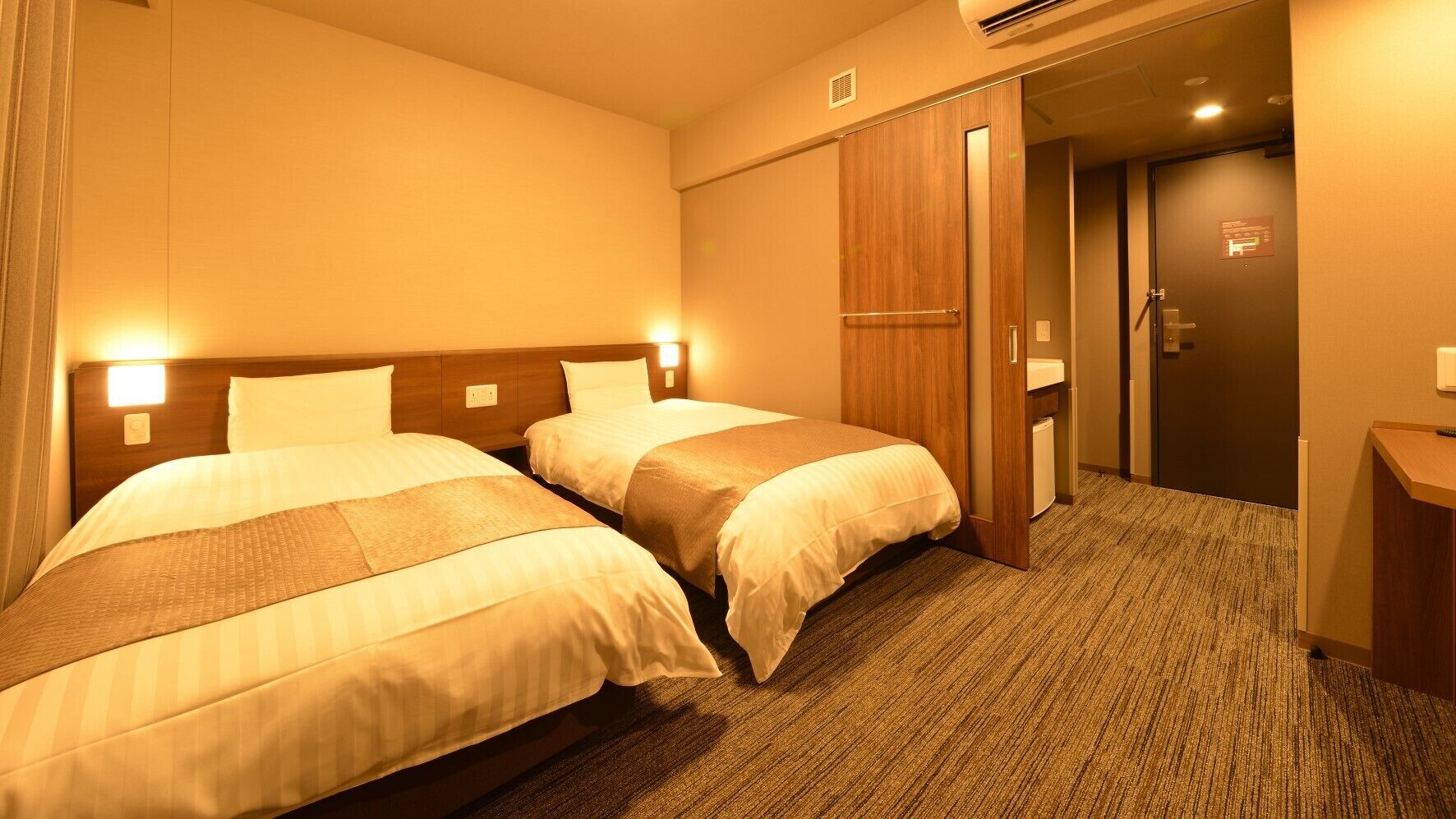 Ein Hotelzimmer mit zwei Betten, einem Holzschrank, einem Schreibtisch und einer Klimaanlage.